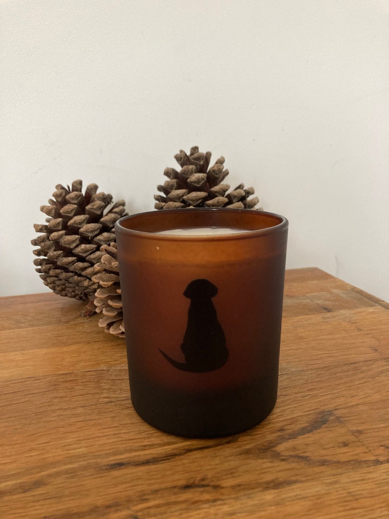 Op de afbeelding: Een bruine, gematteerde glazen kaars met een zwart silhouet van een hond aan de voorkant. De kaars staat op een houten oppervlak met twee dennenappels.