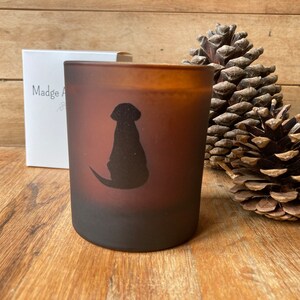 Op de afbeelding: Een bruine, gematteerde glazen kaars met een zwart silhouet van een hond die aan de zijkant zit. De kaars staat op een houten oppervlak met dennenappels.
