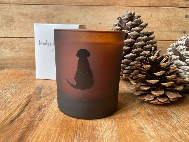Op de afbeelding: Een bruine, gematteerde glazen kaars met een zwart silhouet van een hond die aan de zijkant zit. De kaars staat op een houten oppervlak met dennenappels.