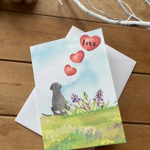 May include: A watercolor greeting card featuring a black dog sitting in a field of flowers. The dog is looking up at three red hearts floating in the sky. The card says "love" in black lettering.