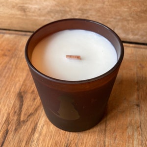 Op de afbeelding: Een bruine glazen kaars met een witte was bovenkant en een houten lont. De kaars heeft een silhouet van een hond aan de zijkant.