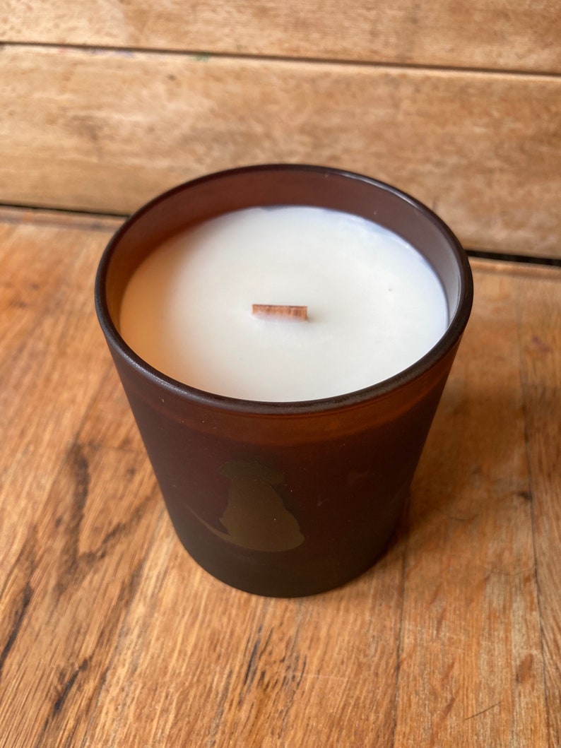 Op de afbeelding: Een bruine glazen kaars met een witte was bovenkant en een houten lont. De kaars heeft een silhouet van een hond aan de zijkant.