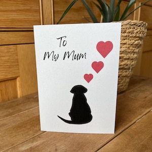 Op de afbeelding: Witte wenskaart met de tekst "To My Mum" en een zwart silhouet van een hond. Drie rode harten zweven boven de hond. De kaart ligt op een houten oppervlak.