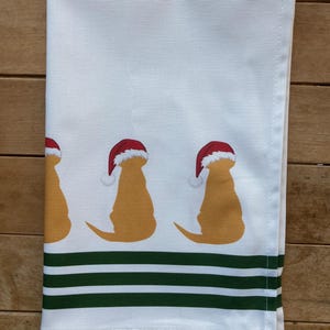 Pode incluir: Toalha de cozinha branca com tr&ecirc;s silhuetas de golden retrievers usando gorros de Papai Noel. A toalha tem uma borda listrada verde e branca. Um design festivo para a &eacute;poca natal&iacute;cia.
