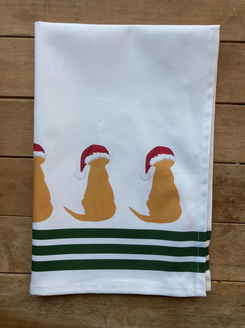 Pode incluir: Toalha de cozinha branca com tr&ecirc;s silhuetas de golden retrievers usando gorros de Papai Noel. A toalha tem uma borda listrada verde e branca. Um design festivo para a &eacute;poca natal&iacute;cia.