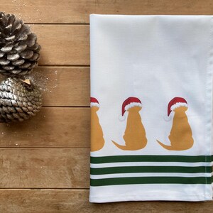 Pode incluir: Toalha de cozinha branca com um padr&atilde;o repetido de silhuetas de golden retrievers usando chap&eacute;us de Papai Noel. A parte inferior da toalha tem tr&ecirc;s listras verdes. Dois pinhas em uma superf&iacute;cie de madeira.
