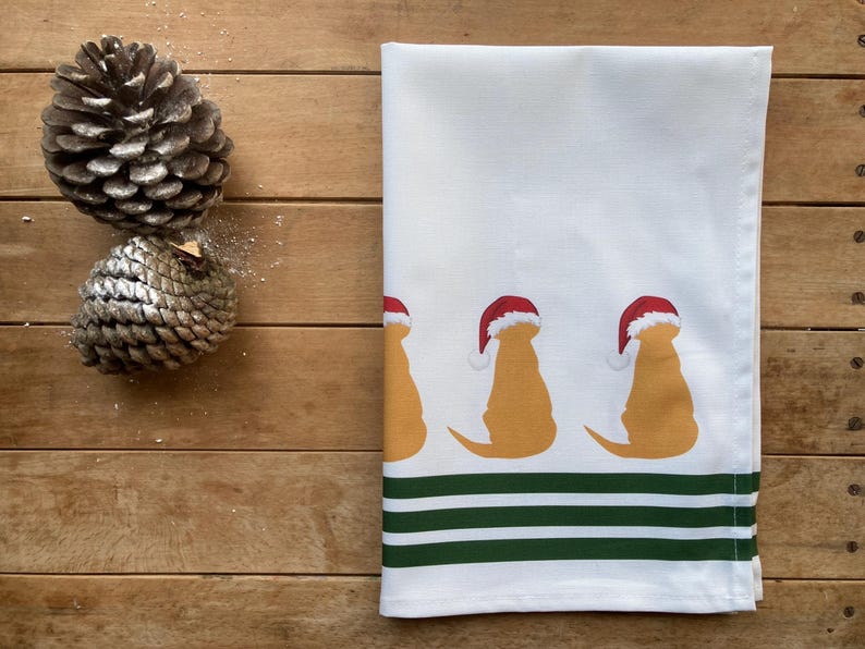 Pode incluir: Toalha de cozinha branca com um padr&atilde;o repetido de silhuetas de golden retrievers usando chap&eacute;us de Papai Noel. A parte inferior da toalha tem tr&ecirc;s listras verdes. Dois pinhas em uma superf&iacute;cie de madeira.