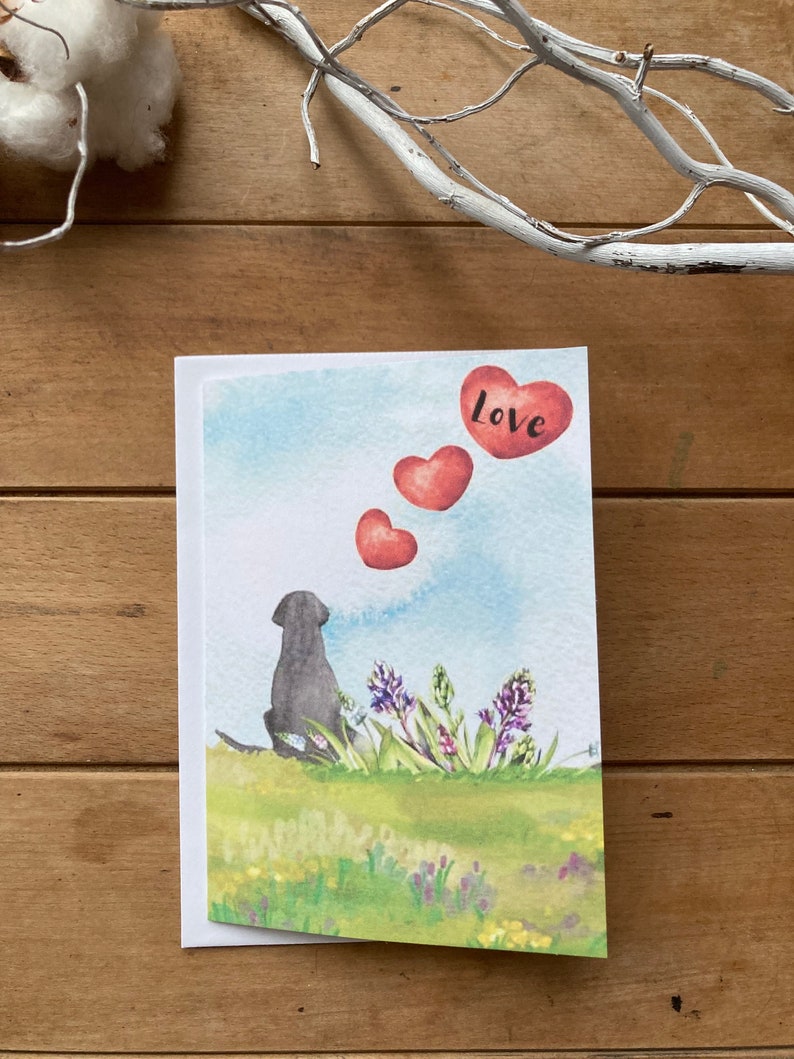 May include: A watercolor greeting card featuring a black dog sitting in a field of flowers, looking up at three red hearts in the sky. The top heart has the word "love" written on it.