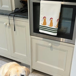 Pode incluir: Toalha de cozinha branca com um design festivo com tr&ecirc;s c&atilde;es castanhos usando chap&eacute;us de Papai Noel, acentuada com listras verdes. A toalha est&aacute; pendurada na porta de um forno em uma cozinha creme.