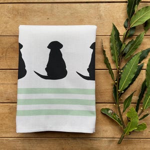 May include: White kitchen towel featuring black dog silhouettes and light green stripes. The towel is folded on a wooden surface, with a sprig of green leaves to the side.