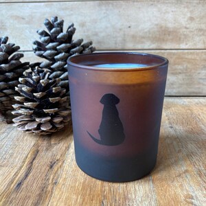 Op de afbeelding: Een bruine, gematteerde glazen kaarshouder met een zwart silhouet van een hond die aan de zijkant zit. De kaars staat op een houten oppervlak met dennenappels.