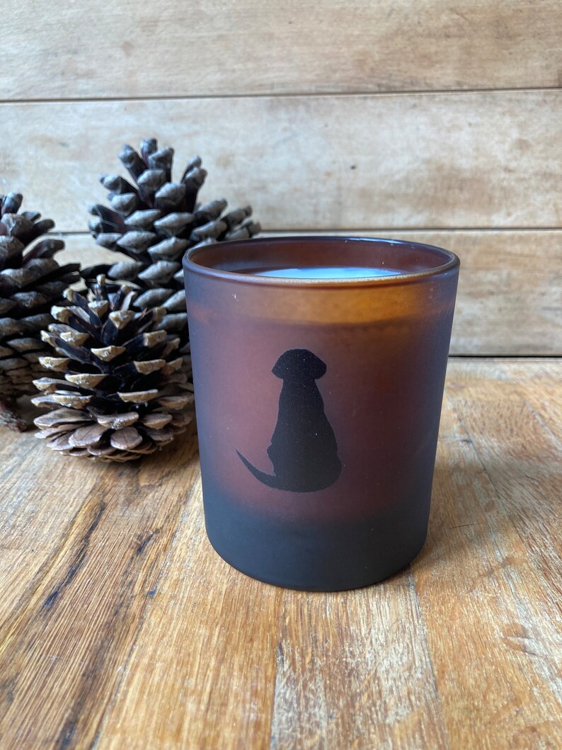 Op de afbeelding: Een bruine, gematteerde glazen kaarshouder met een zwart silhouet van een hond die aan de zijkant zit. De kaars staat op een houten oppervlak met dennenappels.
