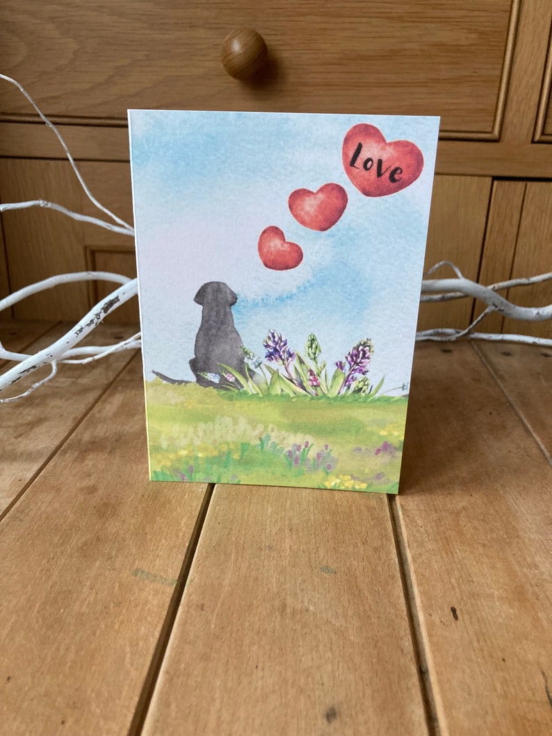 May include: A watercolor greeting card featuring a black dog sitting in a field of flowers. Three red hearts float above the dog, with the word "love" written on the top heart.
