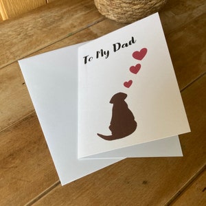Pode incluir: Um cart&atilde;o branco com o texto "To My Dad" em escrita cursiva preta. H&aacute; tr&ecirc;s cora&ccedil;&otilde;es vermelhos acima do texto e uma silhueta marrom de um cachorro olhando para os cora&ccedil;&otilde;es. O cart&atilde;o &eacute; dobrado ao meio e tem um envelope branco.