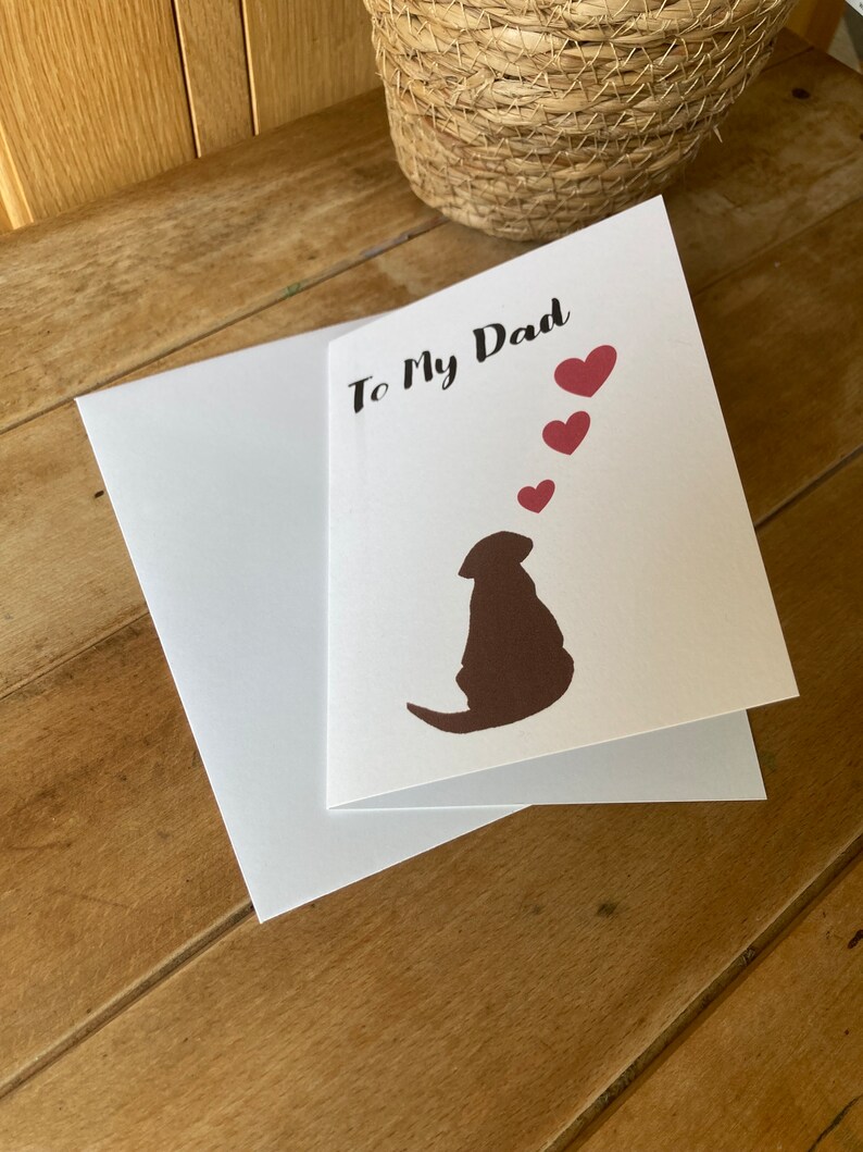 Pode incluir: Um cart&atilde;o branco com o texto "To My Dad" em escrita cursiva preta. H&aacute; tr&ecirc;s cora&ccedil;&otilde;es vermelhos acima do texto e uma silhueta marrom de um cachorro olhando para os cora&ccedil;&otilde;es. O cart&atilde;o &eacute; dobrado ao meio e tem um envelope branco.