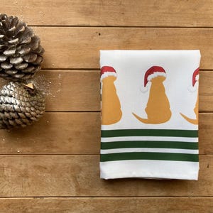 Pode incluir: Uma toalha de cozinha branca dobrada com um padr&atilde;o repetido de silhuetas de c&atilde;es amarelos usando chap&eacute;us de Papai Noel vermelhos. A parte inferior da toalha tem tr&ecirc;s listras verdes e brancas. Dois pinhas est&atilde;o na superf&iacute;cie de madeira.