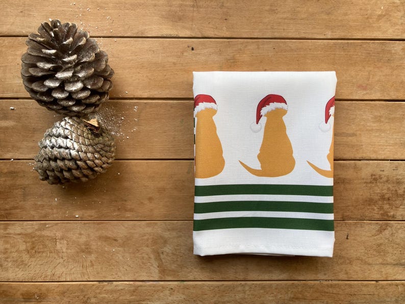 Pode incluir: Uma toalha de cozinha branca dobrada com um padr&atilde;o repetido de silhuetas de c&atilde;es amarelos usando chap&eacute;us de Papai Noel vermelhos. A parte inferior da toalha tem tr&ecirc;s listras verdes e brancas. Dois pinhas est&atilde;o na superf&iacute;cie de madeira.