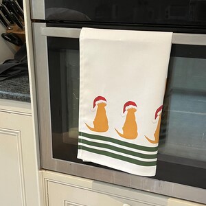 Pode incluir: Toalha de cozinha branca com um padr&atilde;o repetido de silhuetas de golden retrievers usando chap&eacute;us de Papai Noel. A toalha tem listras verdes na parte inferior. A toalha est&aacute; pendurada na porta do forno.