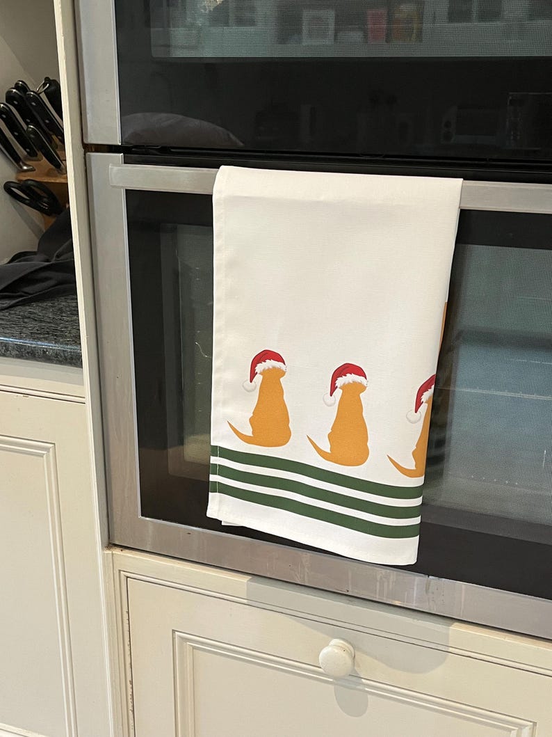 Pode incluir: Toalha de cozinha branca com um padr&atilde;o repetido de silhuetas de golden retrievers usando chap&eacute;us de Papai Noel. A toalha tem listras verdes na parte inferior. A toalha est&aacute; pendurada na porta do forno.