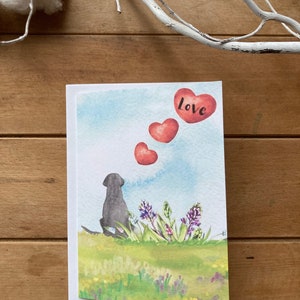 May include: A watercolor greeting card featuring a black dog sitting in a field of flowers, looking up at three red hearts in the sky. The top heart has the word "love" written on it.