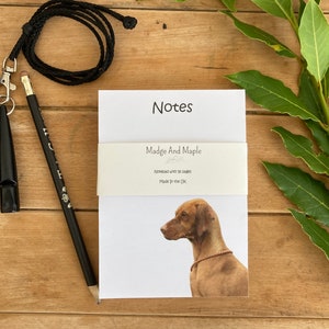 May include: A notepad featuring a dog illustration, a black pencil, and a black whistle with a lanyard. The notepad is branded "Madge And Maple" and has the word "Notes" at the top. Made in the UK.