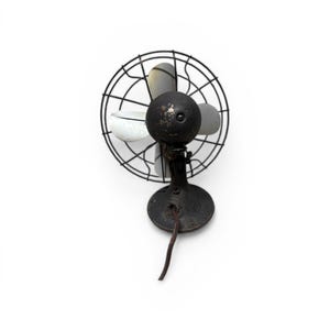 Puede incluir: Un ventilador antiguo de metal negro con una jaula circular y tres aspas. El ventilador tiene una carcasa de motor redonda y una base con un cable de alimentaci&oacute;n. Las aspas son grises y blancas.