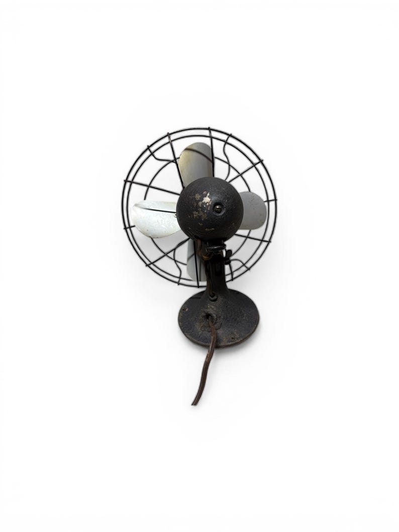 Puede incluir: Un ventilador antiguo de metal negro con una jaula circular y tres aspas. El ventilador tiene una carcasa de motor redonda y una base con un cable de alimentaci&oacute;n. Las aspas son grises y blancas.