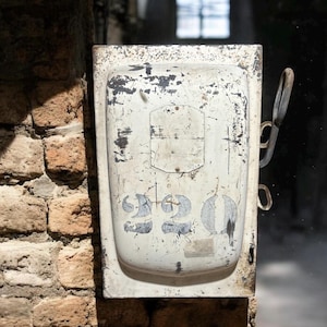 May include: A weathered, white metal electrical box with the number "320" visible. The box is mounted on a brick wall and has a handle on the side. The paint is chipped and worn, giving it a vintage appearance.