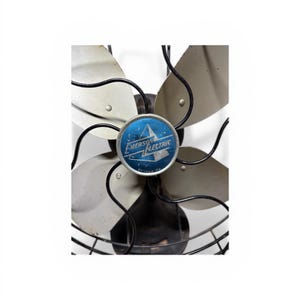 Puede incluir: Ventilador antiguo Emerson Electric con cuatro aspas plateadas y una jaula de alambre negra. El centro del ventilador presenta un emblema azul con las palabras "Emerson Electric" y un dise&ntilde;o de estrella. Una pieza de decoraci&oacute;n cl&aacute;sica.