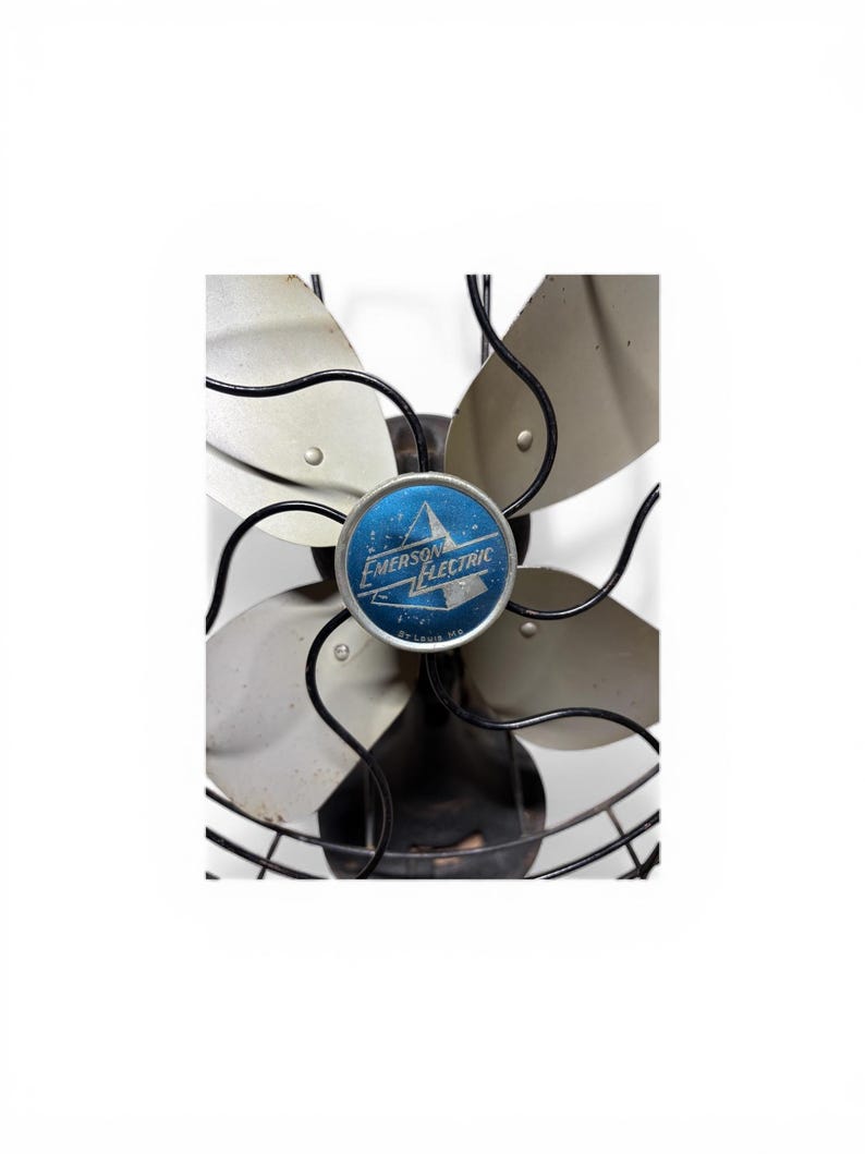 Puede incluir: Ventilador antiguo Emerson Electric con cuatro aspas plateadas y una jaula de alambre negra. El centro del ventilador presenta un emblema azul con las palabras "Emerson Electric" y un dise&ntilde;o de estrella. Una pieza de decoraci&oacute;n cl&aacute;sica.