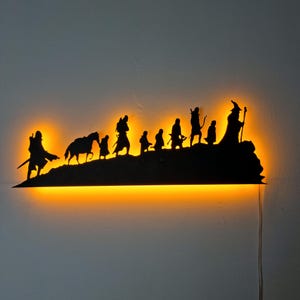 May include: A black metal wall art piece depicting silhouettes of figures walking across a hill, with a warm orange glow emanating from behind. The figures include a wizard, archer, and others, creating a fantasy theme. The piece is designed to be mounted on a wall.