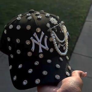 May include: Black baseball cap adorned with numerous sparkling rhinestones and a white "NY" logo. The cap features a pearl and chain embellishment, adding a touch of elegance to the sporty design. A stylish accessory.