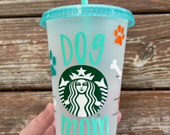 May include: A frosted, clear plastic reusable cup with a teal lid and teal straw. The cup has a green Starbucks logo and the words "Dog Mom" written in teal with paw prints and bone designs around the cup.