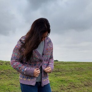 Op de afbeelding: Een lavendelkleurig vest met een kleurrijk, gestructureerd patroon. De trui met knopen heeft een V-hals en geribde manchetten. Het ontwerp bevat verschillende kleuren, waaronder blauw, geel en rood. Het vest wordt over een grijze top gedragen.