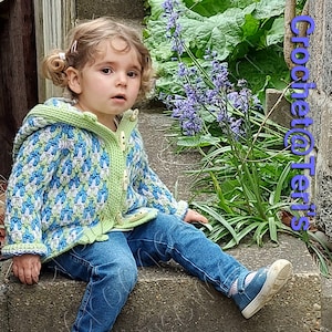 Peut inclure: Un jeune enfant portant une veste à capuche en crochet vert, bleu et blanc avec des boutons. L'enfant est assis sur un ensemble de marches en béton avec une plante verte en arrière-plan.