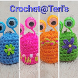 May include: Four handmade crochet monster keyrings in vibrant colours. Each has large, plastic googly eyes and a unique design. The keyrings are blue with a green flower, pink with embroidered flowers, orange with a green and yellow pattern, and green with a purple flower. The text "Crochet@Teri's" is visible.
