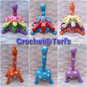 May include: A set of five crocheted dinosaur toys. The dinosaurs are stacked on top of each other, with the largest dinosaur at the bottom and the smallest dinosaur at the top. The dinosaurs are all different colours, including purple, blue, orange, and pink. Each dinosaur has a white flower pattern on its body.