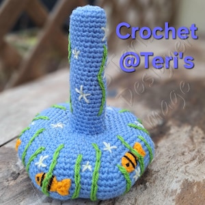 Peut inclure: Une sc&egrave;ne sous-marine en crochet bleu et vert avec des fleurs blanches et des poissons orange. La pi&egrave;ce en crochet a la forme d'un vase avec un long col et une base large. Le texte "Crochet @ Teri's" est visible sur l'image.