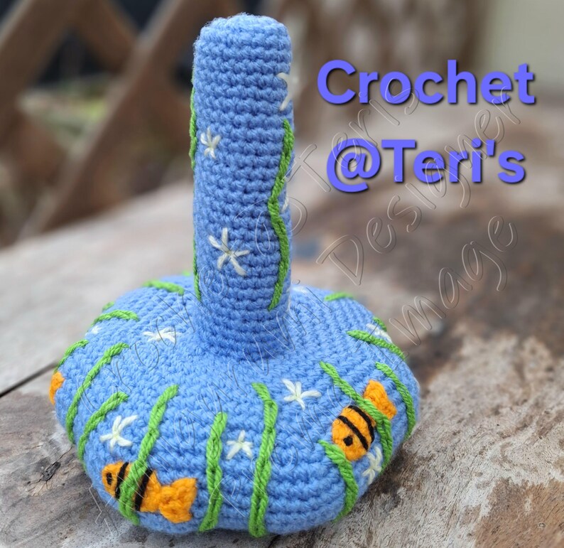 Peut inclure: Une sc&egrave;ne sous-marine en crochet bleu et vert avec des fleurs blanches et des poissons orange. La pi&egrave;ce en crochet a la forme d'un vase avec un long col et une base large. Le texte "Crochet @ Teri's" est visible sur l'image.