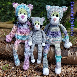 May include: Three crocheted cat toys sitting on a log. The largest cat is purple, blue, and green. The middle cat is grey. The smallest cat is blue, purple, and green.