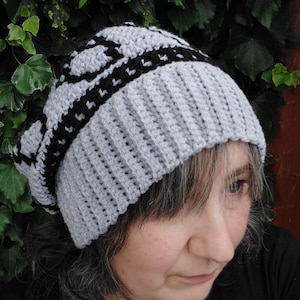 May include: A gray and black crocheted beanie with a geometric pattern. The beanie is worn by a person with gray hair.
