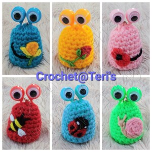 May include: Six crocheted amigurumi toys with googly eyes. Each toy is a different color and has a different design. The designs include a bee, a ladybug, a snail, a rose, a flower, and a ladybug. Crochet@Teri's is written on the image.