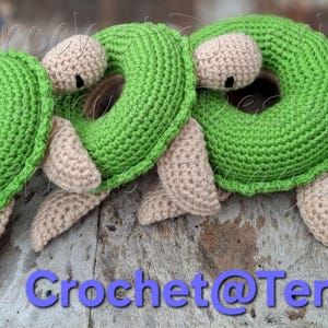 Peut inclure: Trois jouets en crochet en forme de tortue verte avec des visages et des membres beiges. Les tortues sont en forme de beigne avec un trou au centre. Les jouets sont sur une surface en bois.