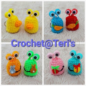 May include: Six crocheted amigurumi keychains with googly eyes. Each keychain features a different color and design, including a yellow keychain with a carrot, a blue keychain with a carrot, a red keychain with a carrot, an orange keychain with a bunny, a green keychain with a bunny, and a pink keychain with a gift.