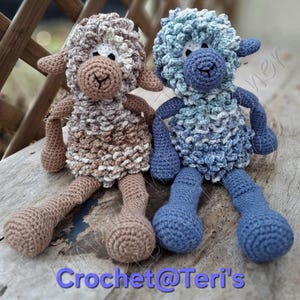 May include: Two handmade crochet sheep dolls. One is brown and cream, the other is blue and white. They have fluffy wool-like bodies, long legs, and small arms. The dolls are sitting on a wooden surface. The text "Crochet@Teri's" is at the bottom.