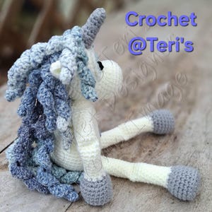 May include: A crocheted unicorn doll in cream, blue, and grey. The unicorn has a grey horn, blue and grey mane, and grey hooves. The text "Crochet @Teri's" is visible in the upper right corner.