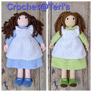 May include: Two handmade crochet dolls. One doll wears a blue dress with a white apron and blue shoes. The other doll wears a green dress with a white apron and green shoes. Both dolls have brown curly hair and are displayed on a wooden surface. The text "Crochet @ Teri's" is at the top.