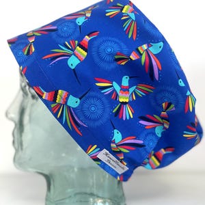 May include: A blue scrub cap featuring a repeating pattern of colourful hummingbirds with wings in shades of red, yellow, green, and purple, accented by dark blue circular starburst designs. A white fabric tag reads Howellmade Devon.