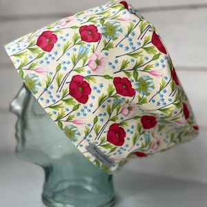 May include: A white scrub cap with a floral pattern featuring pink and red poppies, blue flowers, and green leaves. The cap has a tie at the back.