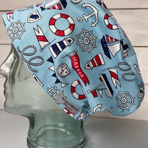 May include: A light blue surgical scrub hat with a nautical theme. The hat features a variety of nautical symbols, including anchors, ships, life preservers, and steering wheels. The text "Harbor" is printed on the hat.